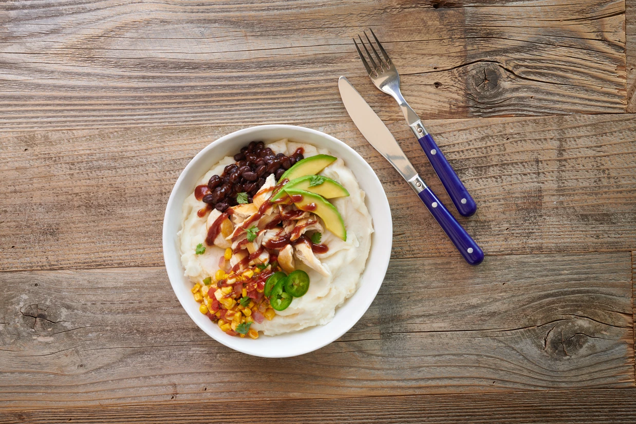 A bowl of mashed potatoes topped with sliced avocado, corn, black beans, jalapeños, shredded chicken, and barbecue sauce sits on a wooden table—an inventive twist reminiscent of Thai food—next to a fork and knife with blue handles.
